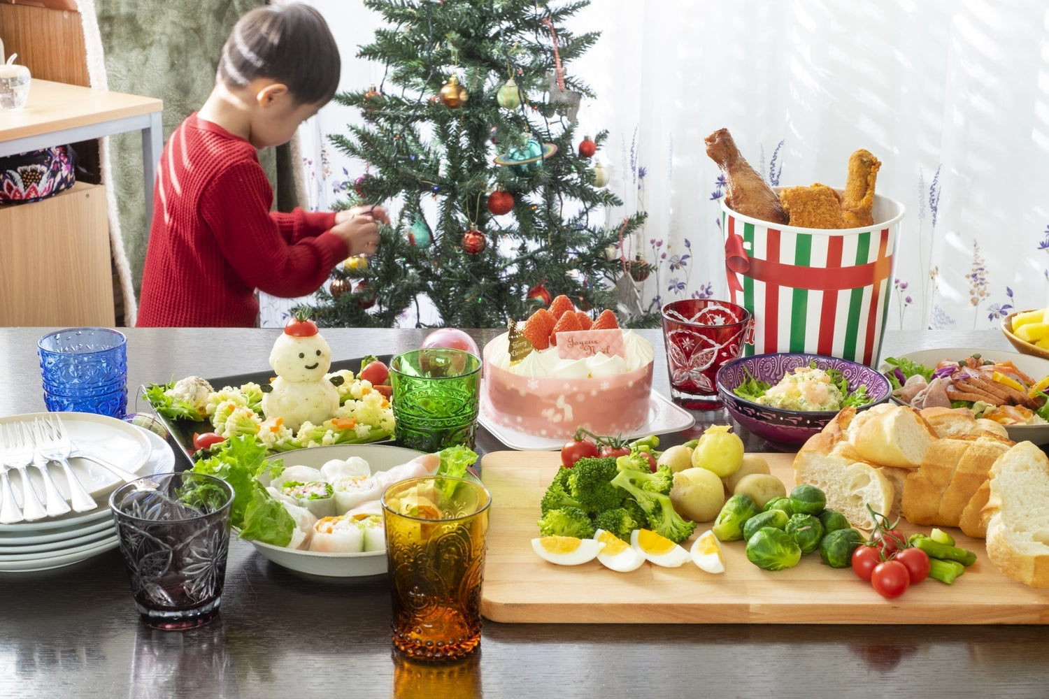Enfant décorant un sapin de Noël derrière une table chargée de nourriture dont un fraisier et un sceau de poulet frit