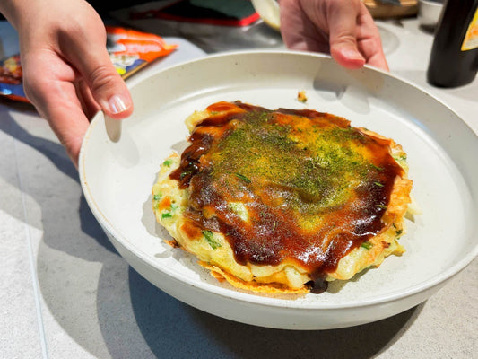 Mains tenant une assiette avec un okonomiyaki recouvert de sauce et d'aonori