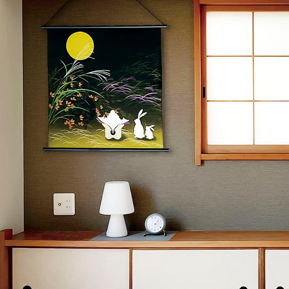 Decorative wall hanging with two white rabbits under a yellow moon on a dark background, placed above a wooden desk with a lamp and clock.